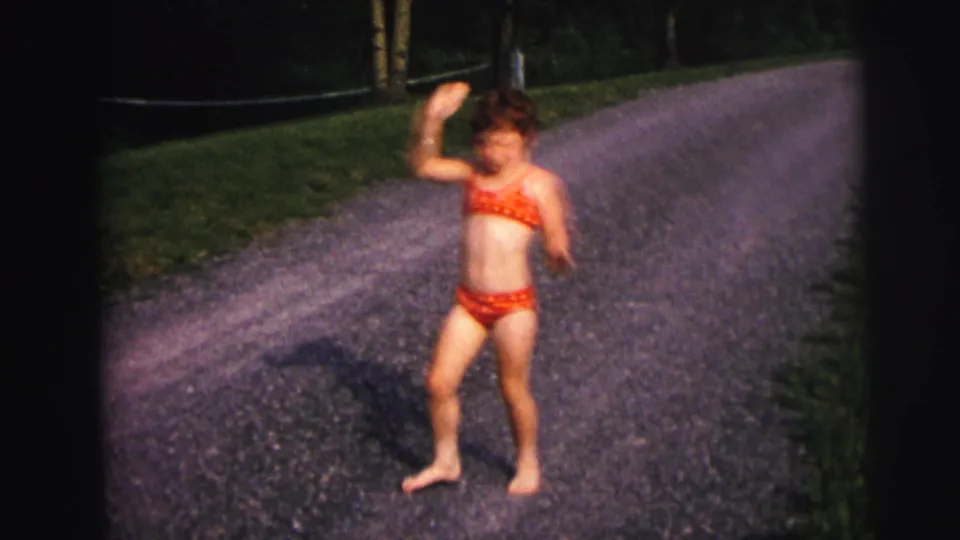 A child in red swimwear stands on a gravel lakeside road, with grass, trees, and water behind them in 1970s home-movie footage.