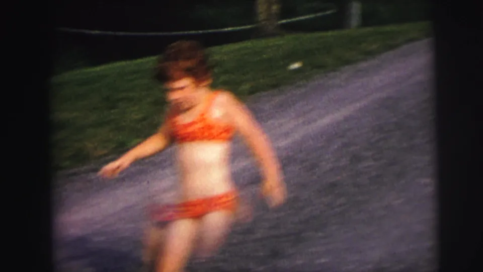 Motion-blurred 8mm frame of a child in red swimwear moving beside a gravel lakeside road with grass and water nearby.