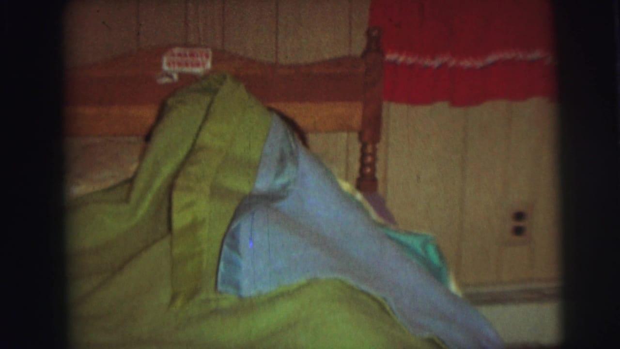 A 1970s 8mm home-movie frame shows green and pale blue blankets on a wooden bed, with wall paneling and a red curtain behind them.