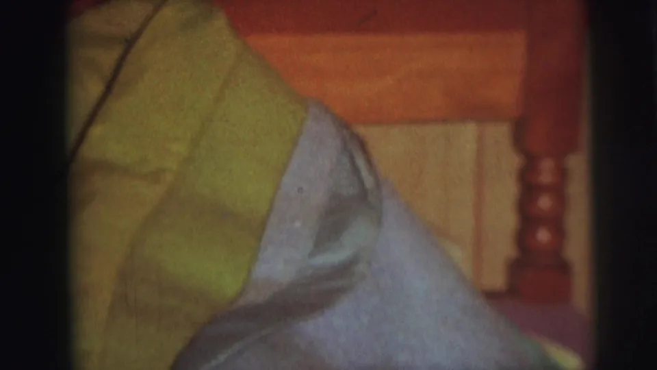 The blankets fill the foreground while a wooden headboard, bedpost, and paneled wall remain visible in the soft 8mm home-movie frame.