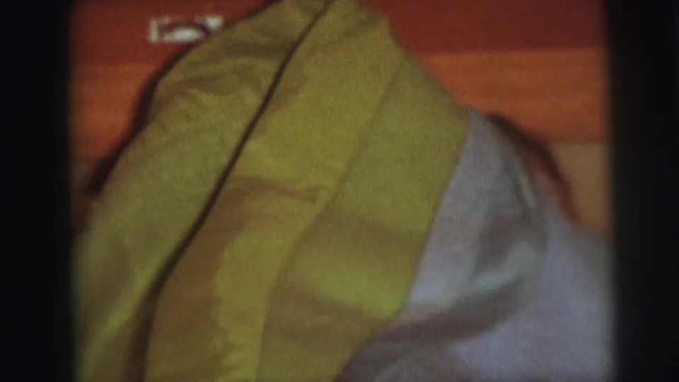 A close home-movie view shows bunched green and pale blue blankets on a wooden bed, with a small label-like object near the headboard.