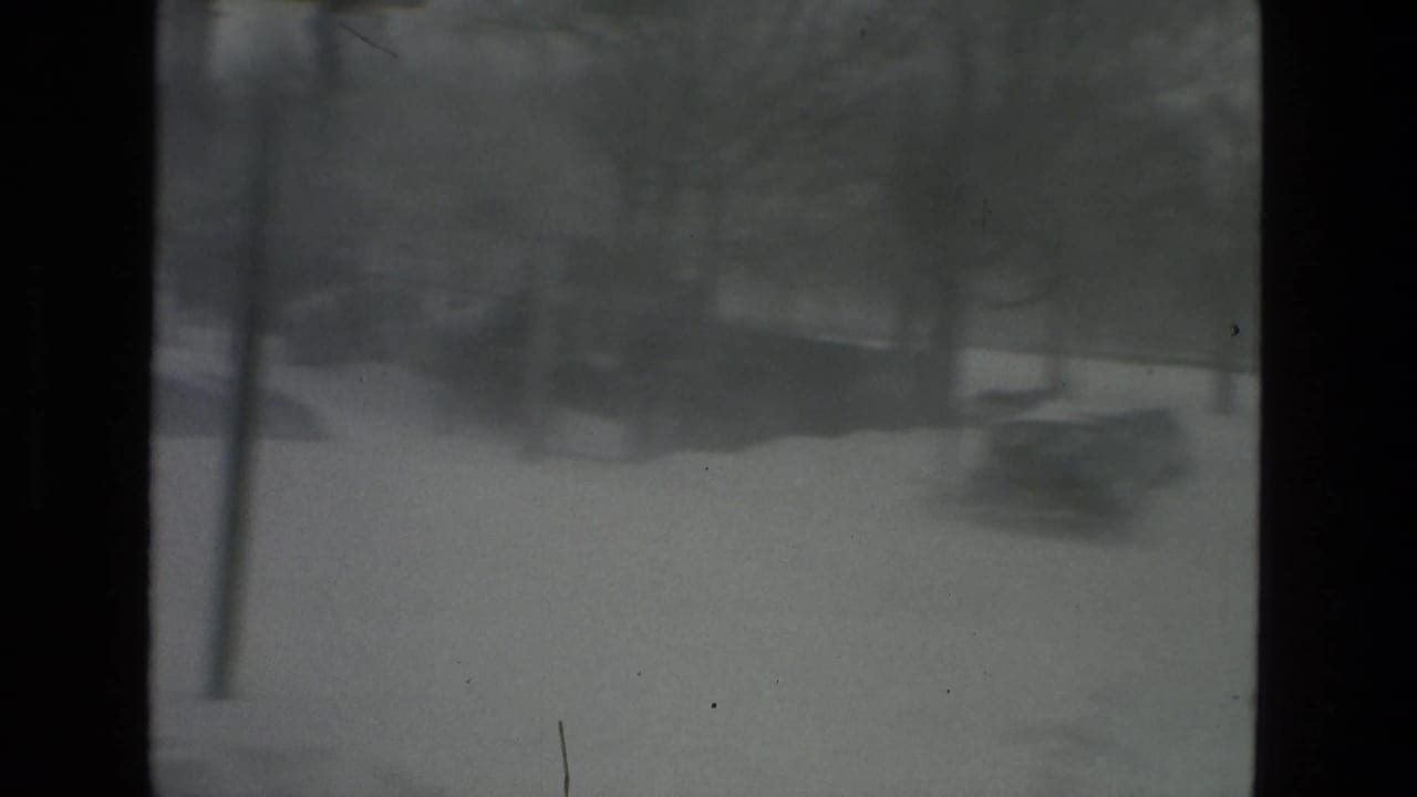 Soft, grainy 8mm view of a snow-covered urban street during a heavy storm, with dark vertical frame edges and indistinct trees or roadside shapes in the distance.