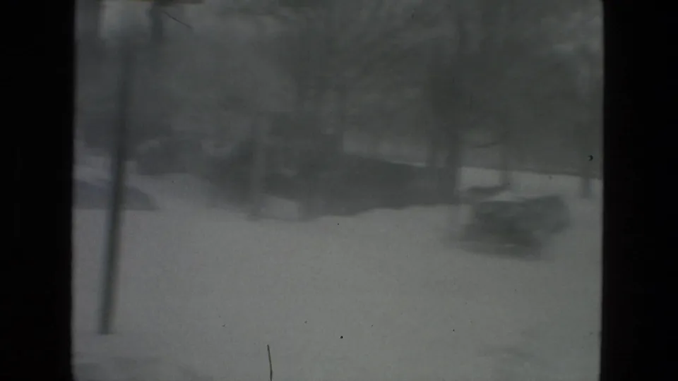 Soft, grainy 8mm view of a snow-covered urban street during a heavy storm, with dark vertical frame edges and indistinct trees or roadside shapes in the distance.