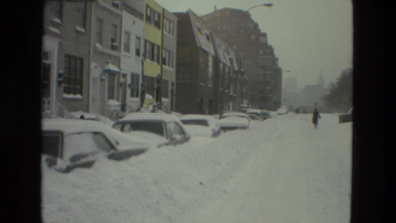 Snow-covered city street with parked cars partly buried beside row houses.