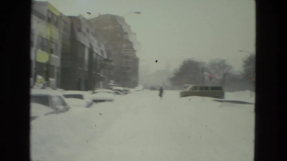 A snowbound urban block shows parked cars almost covered by drifts, multi-story buildings along the left side, and a small pedestrian figure crossing through the storm haze.
