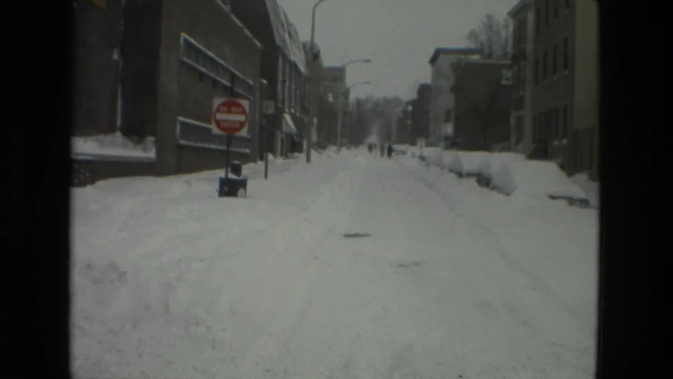 A quiet snow-covered city street stretches past brick buildings and a visible Do Not Enter sign, with small figures walking far down the road under gray winter light.