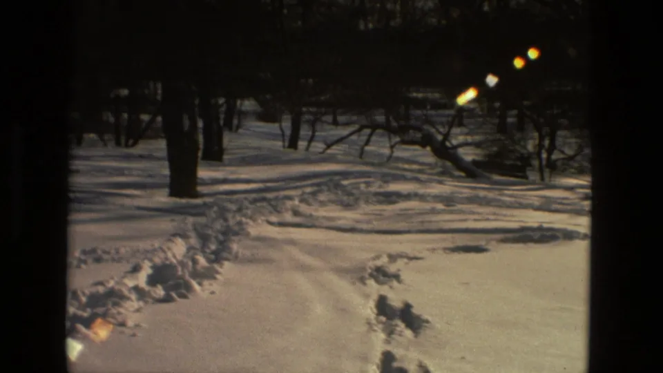 Vintage 8mm winter frame showing footprints or tracks in snow, bare trees, heavy shadows, and small flare highlights.