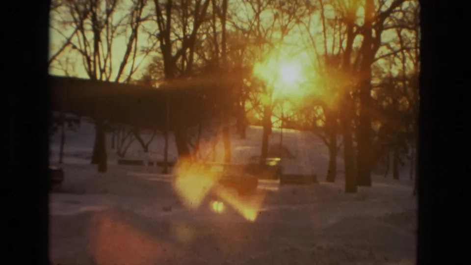 1978 8mm home-movie frame showing low golden sunlight through bare trees above a snowy outdoor landscape, with flare and soft focus.
