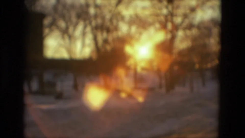 Soft 8mm frame of a snowy outdoor area with bare trees, warm sunlight, and pronounced lens flare during the zoom movement.