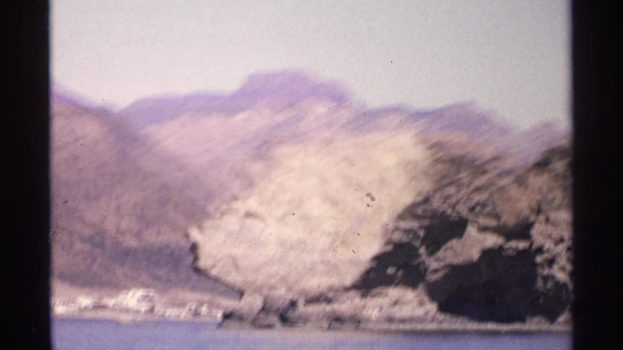 1975 Guaymas, Mexico 8mm frame showing blue water, pale coastal cliffs, dark rocks, and hazy mountains beyond the bay.