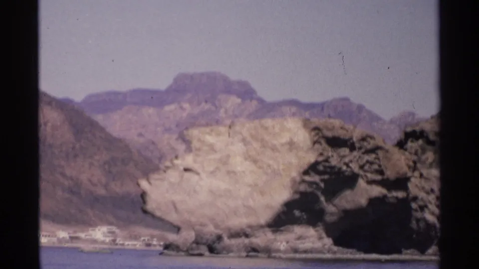 1975 Guaymas, Mexico 8mm view of blue water beneath pale cliffs, dark rocky shore, distant buildings, and hazy mountains.