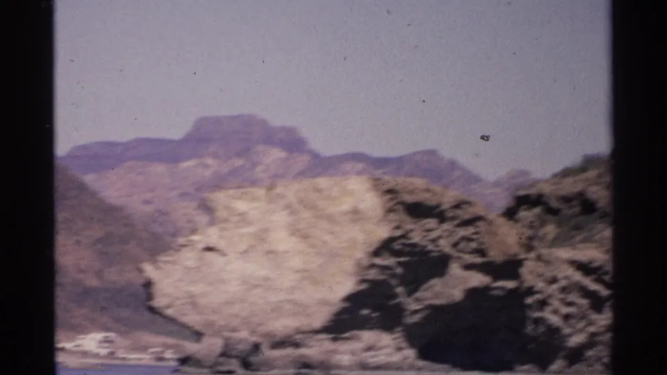 1975 Guaymas, Mexico frame showing pale cliff strata, dark rocks, blue water, and soft hazy mountain ranges.
