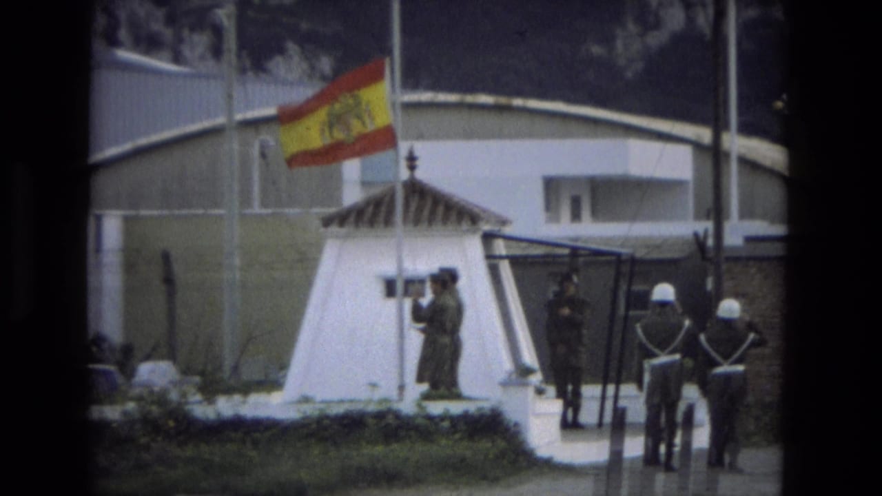 Soldier lowers a Spanish flag near a white sentry post as guards stand nearby.