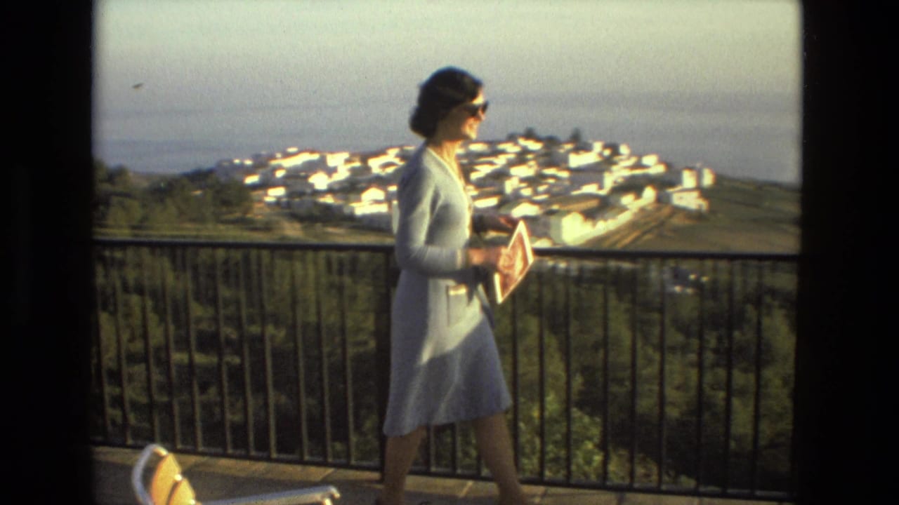 Woman in a light dress walks on a balcony above a white coastal town.