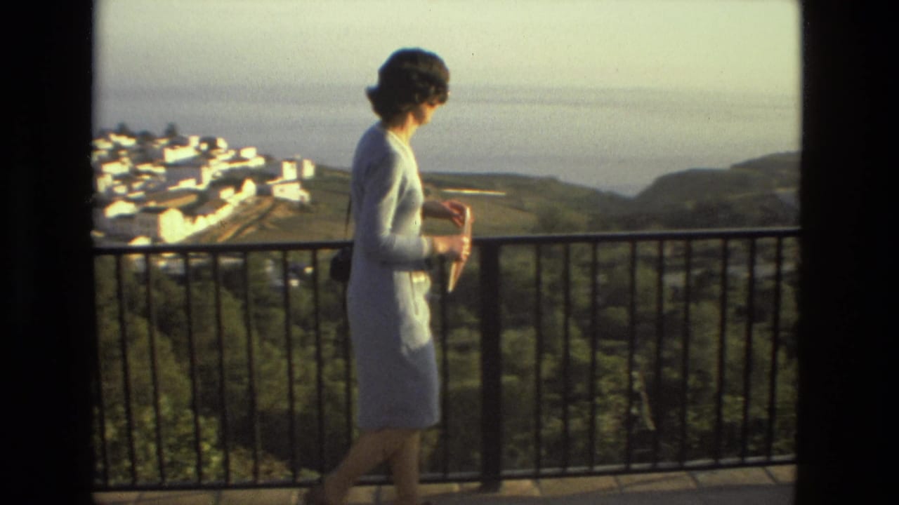 Woman walks past a balcony railing with hills, white buildings, and sea in the background.