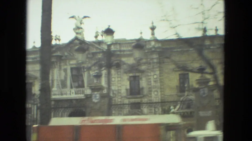 An ornate historic building in Andalusia appears through bare branches, with decorative roof details, a lamp, and a red-and-white vehicle below.