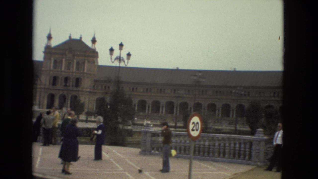 Pedestrians stand near a 20 km/h sign with an arched historic building behind them.