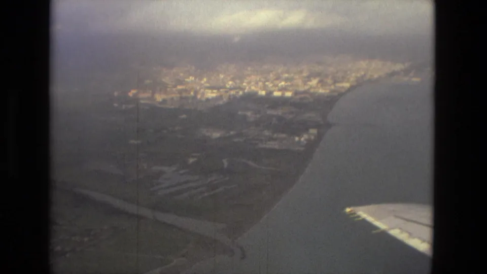 A hazy airplane-window view shows a coastal city, shoreline, water, and part of the aircraft wing during the travel sequence.