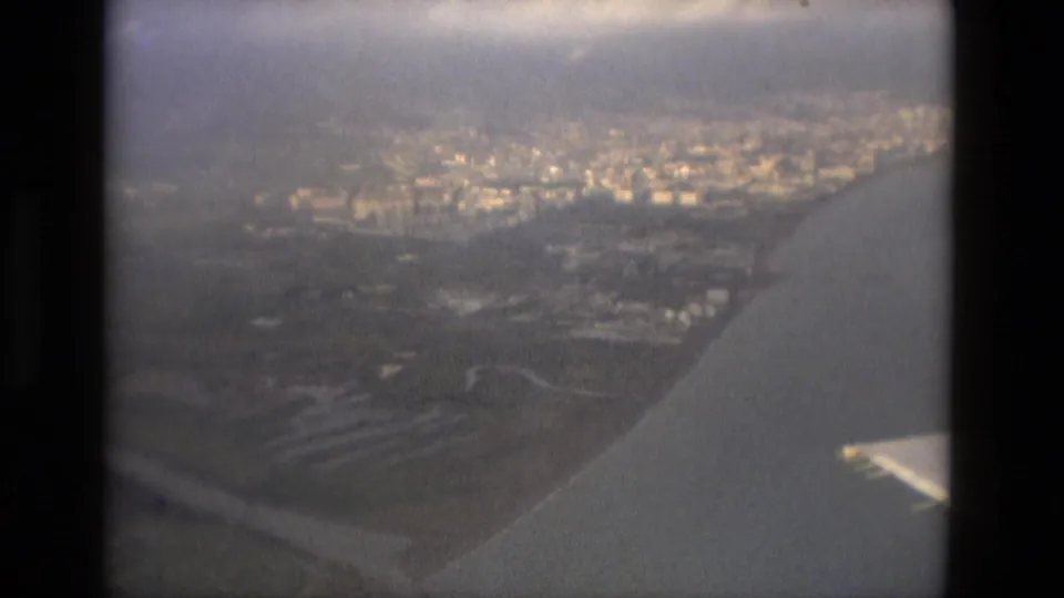 The final frame continues the hazy passenger-window view over a coastal city, with water and the airplane wing visible at lower right.