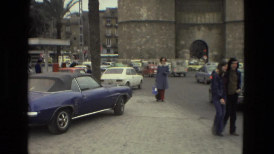Two pedestrians walk near a blue convertible as vintage cars and a stone tower fill the Andalusia street background.