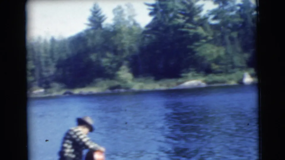 1956 Traverse City home-movie frame showing a man in a plaid shirt and hat near a calm wooded lake.