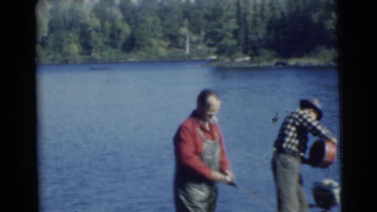 Two men by a lake, one holding a fishing rod and another bending near a small motor.