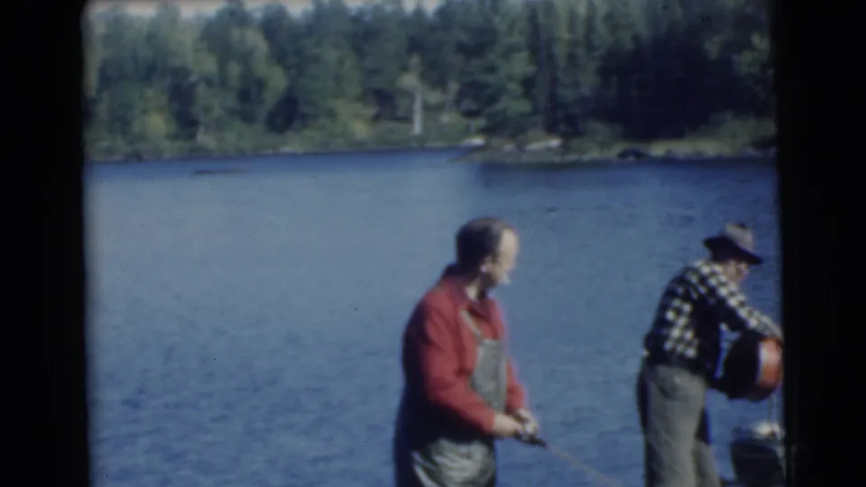 1956 home-movie frame from Traverse City showing two men fishing beside calm water and a tree-lined shore.