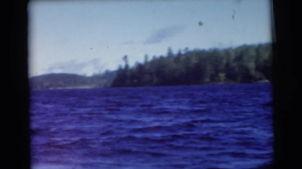 1956 8mm home-movie view of blue water near Traverse City, with a wooded shoreline and pale sky beyond.
