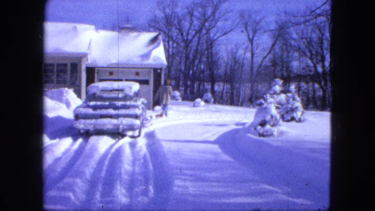 Snowy driveway with a parked car, garage, bare trees, and a person near the yard.