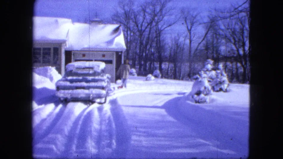 1963 Duluth home-movie frame showing a snow-covered car by a garage as a bundled person stands near the snowy yard.
