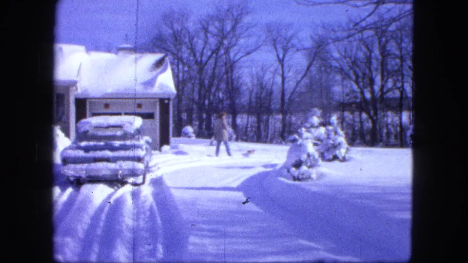 1963 Duluth home-movie frame showing a bundled person and small dog in a snow-covered suburban driveway area.