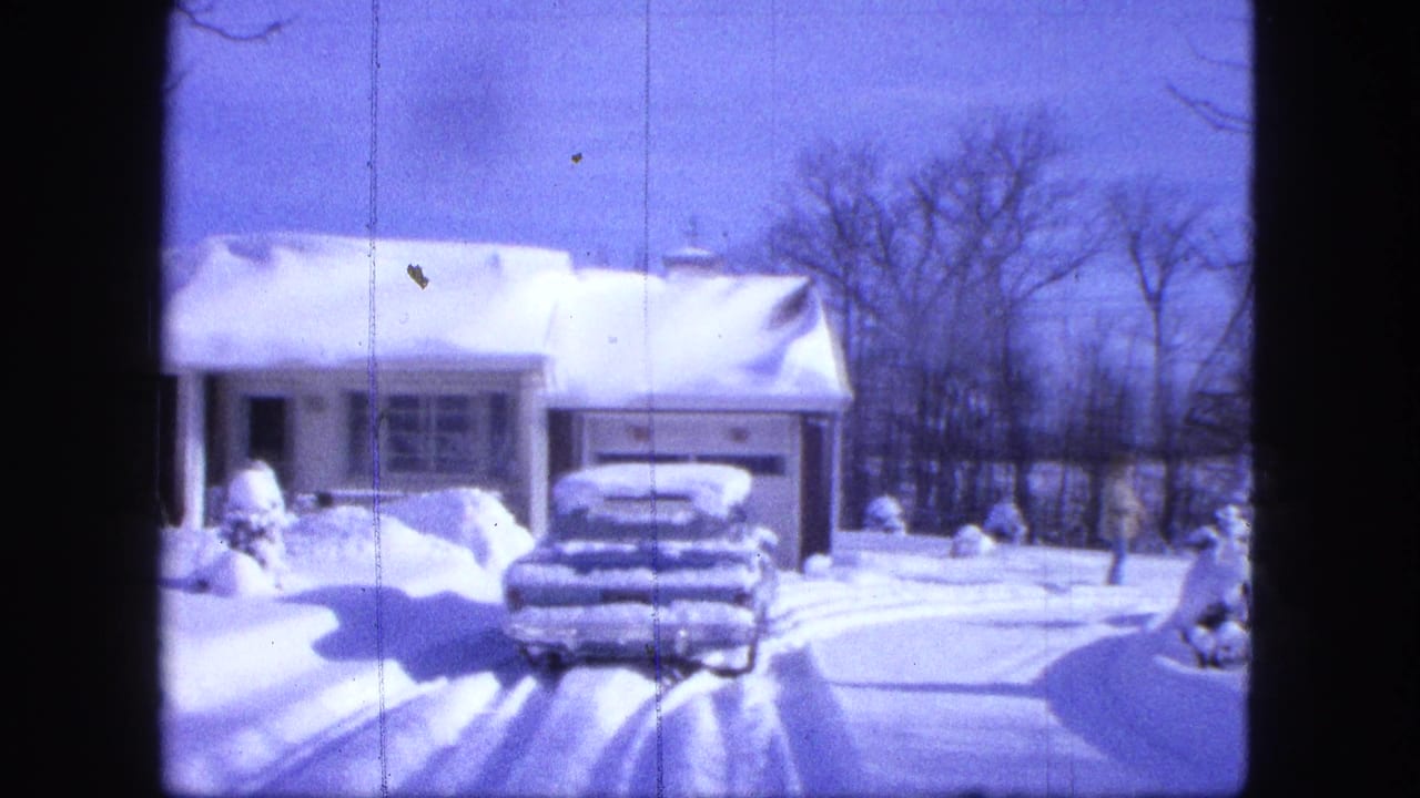 1963 Duluth home-movie frame showing a snow-covered house, buried car, driveway tracks, and bare trees under blue sky.