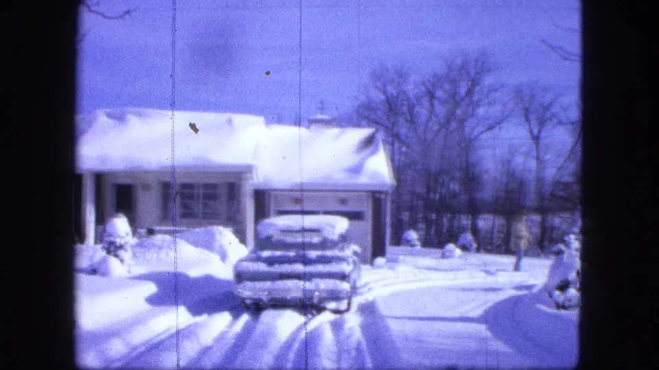 1963 Duluth home-movie frame showing a snow-covered house, buried car, driveway tracks, and bare trees under blue sky.