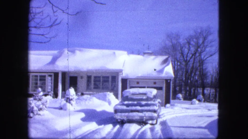1963 Duluth winter frame with a single-story house, snow-covered roof, car in the driveway, and bare trees beyond the yard.