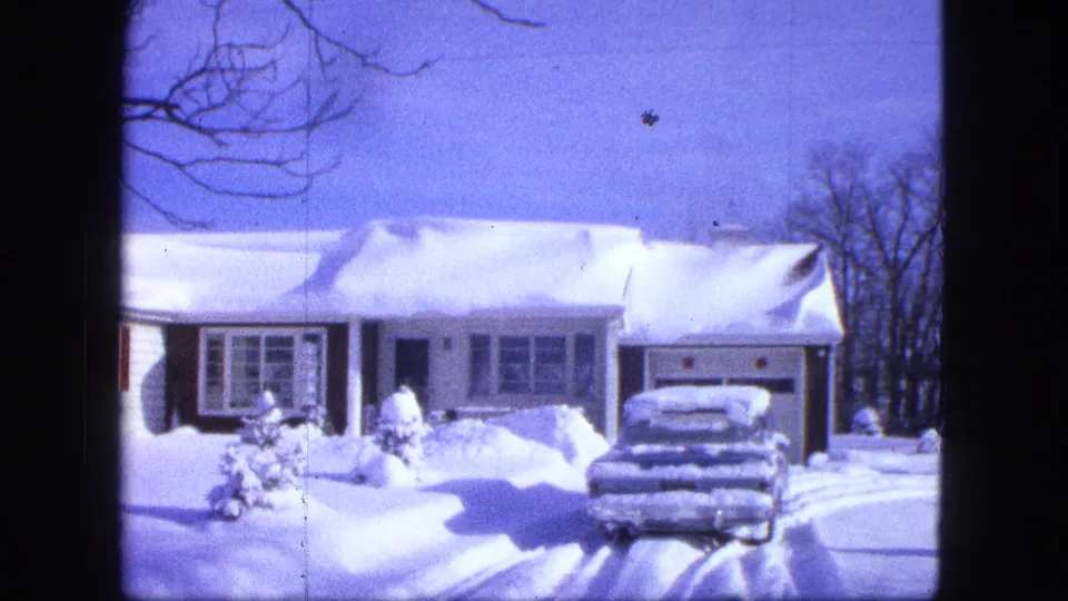 1963 Duluth home-movie view of the snowy house front, buried car, red shutters, and a bundled person near the walkway.