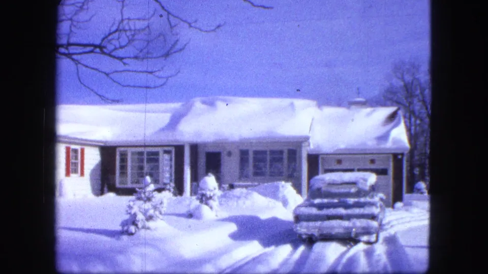 1963 Duluth frame showing a residential house under deep snow, a parked car, driveway tracks, bare branches, and a small bundled figure.