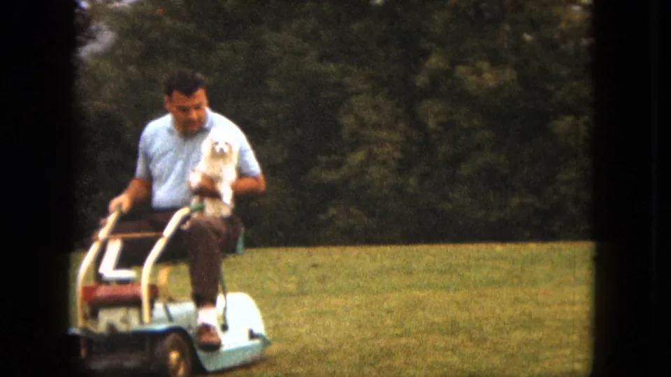In 1963 Duluth home-movie footage, a man in a light blue shirt rides a small lawn mower across grass while holding a small white dog.