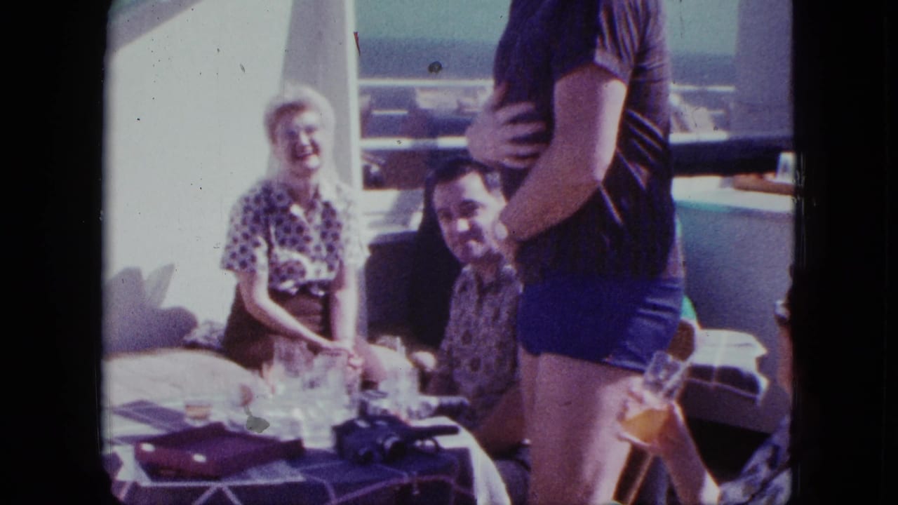 1963 Duluth home-movie frame of adults laughing around a deck table as a man in blue swim trunks stands nearby.