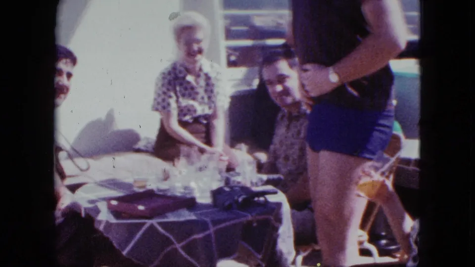 1963 Duluth deck scene with seated adults smiling as a man in swim trunks lifts his shirt beside the table.