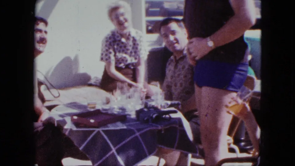 1963 Duluth 8mm frame showing a man in blue swim trunks beside laughing adults at a sunlit deck table.