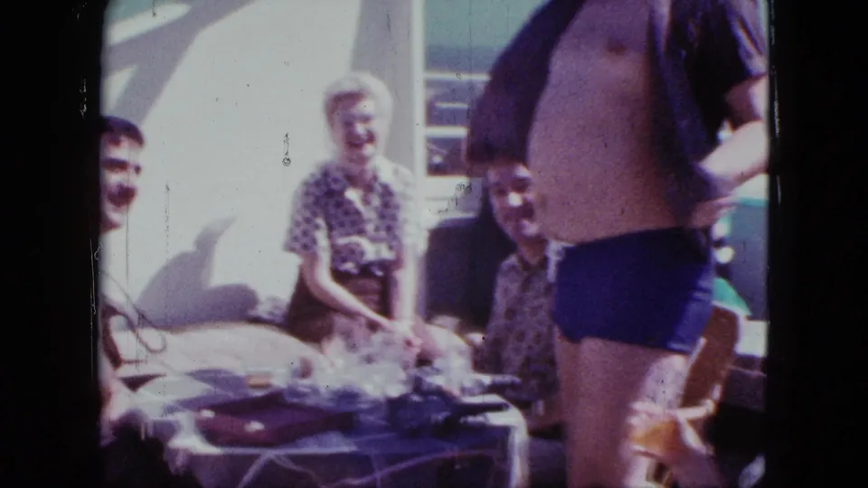 1963 Duluth home-movie still of a man revealing his stomach as seated family members laugh on a deck.