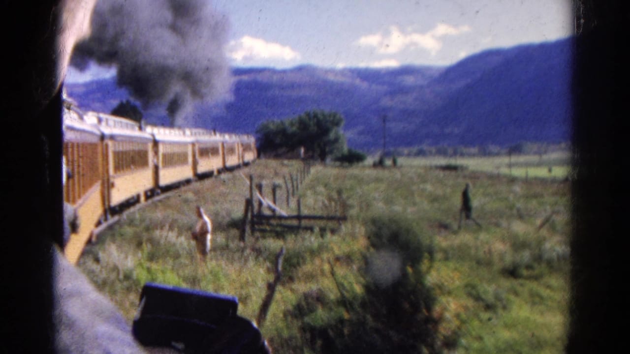 1967 8mm frame of a yellow steam train seen from a passenger-side viewpoint, with smoke, fencing, grass, and mountains beyond.