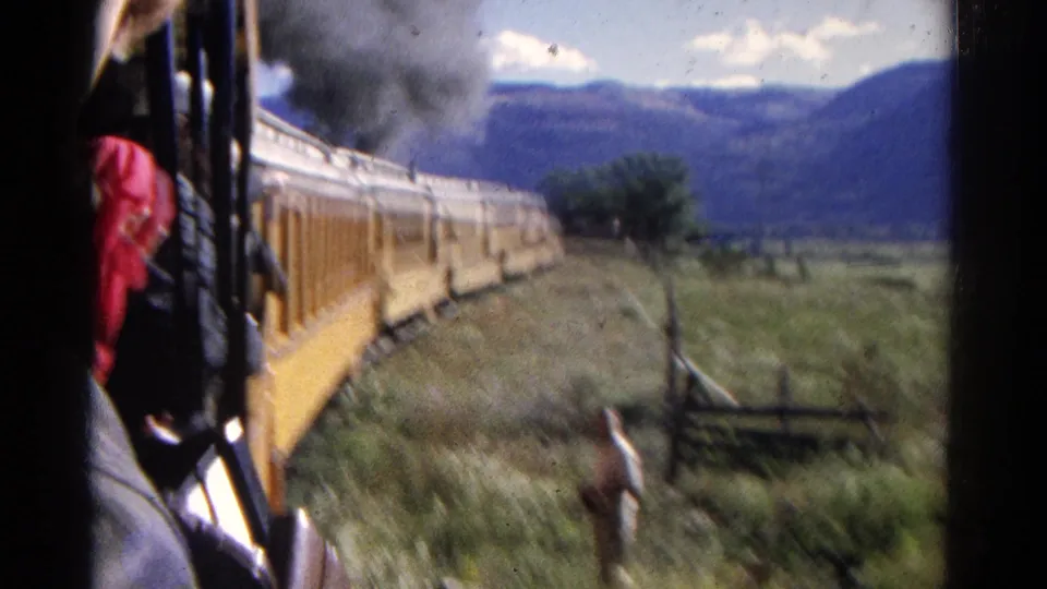 A person is partly visible near the window as the yellow train curves through grassy mountain scenery, with smoke rising ahead.