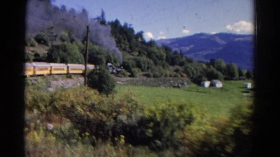 Soft 8mm view of a yellow steam train crossing a green hillside, with dark smoke, trees, utility poles, and mountains in the background.