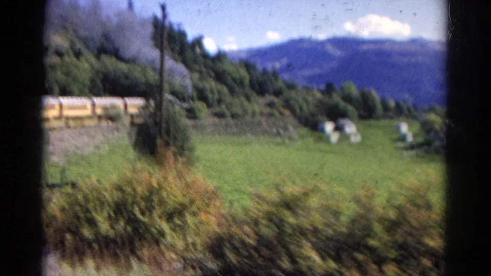 Handheld 8mm frame of the train receding through green rural terrain, with smoke, trees, poles, hay bales, and blue mountains visible.