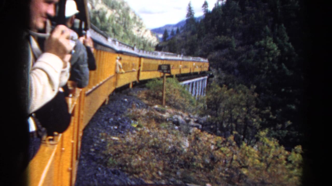 1967 8mm frame of a yellow mountain train rounding a curve, seen from an open car with a passenger holding a camera nearby.