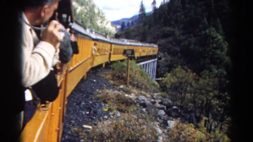 Yellow passenger train seen from an open car as it bends across a trestle through rocky, forested mountain terrain.