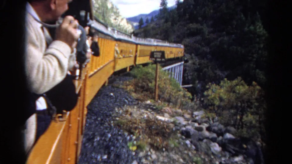 A passenger in a light sweater holds a camera while the yellow train continues along a rocky mountain route.