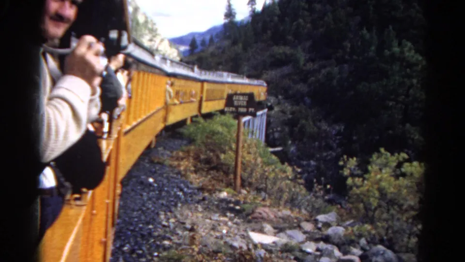 Open-car view of yellow train cars curving past a trackside sign in a wooded mountain setting.