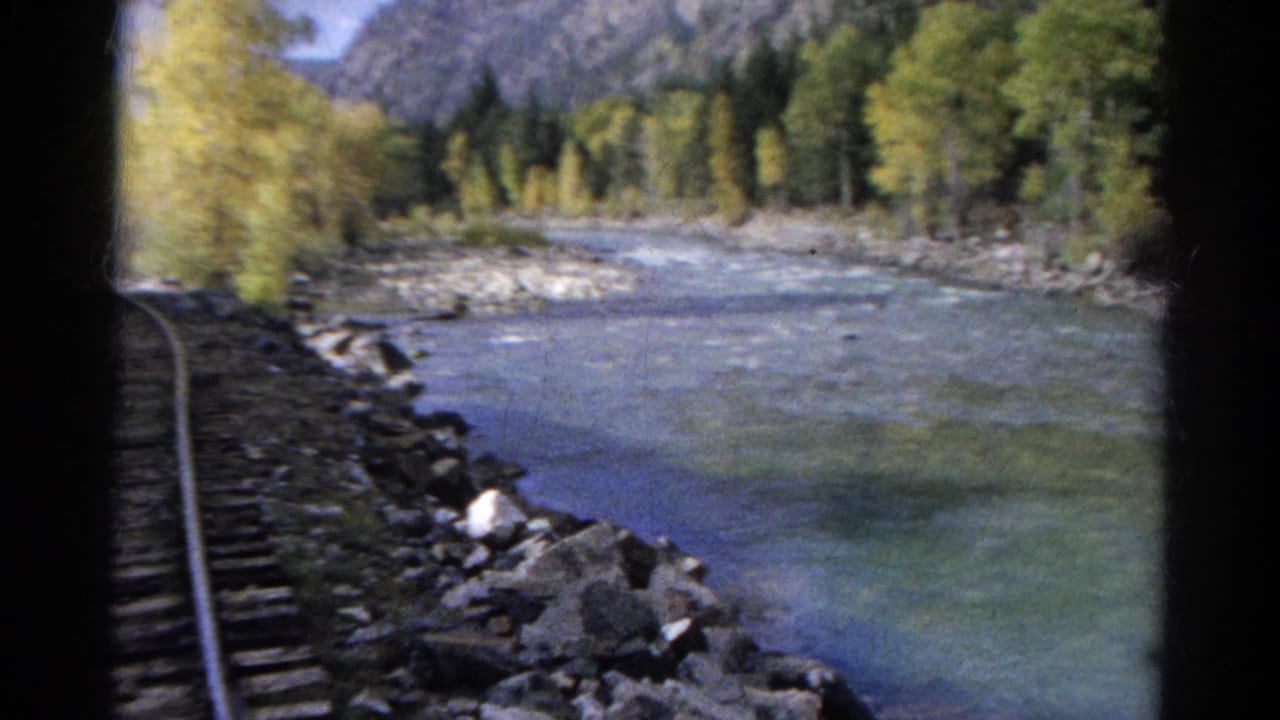A train-window view shows railroad tracks running along a rocky river with autumn trees and mountains beyond.